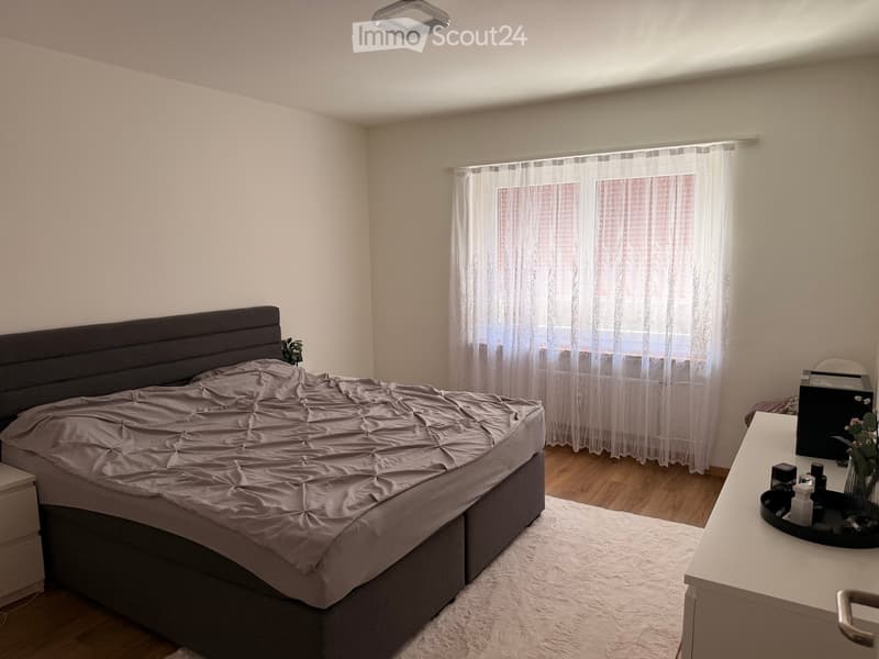 Modern bedroom with a large bed, nightstand, mirror, and a small table with a vase. The room features wooden flooring and large windows with sheer curtains.