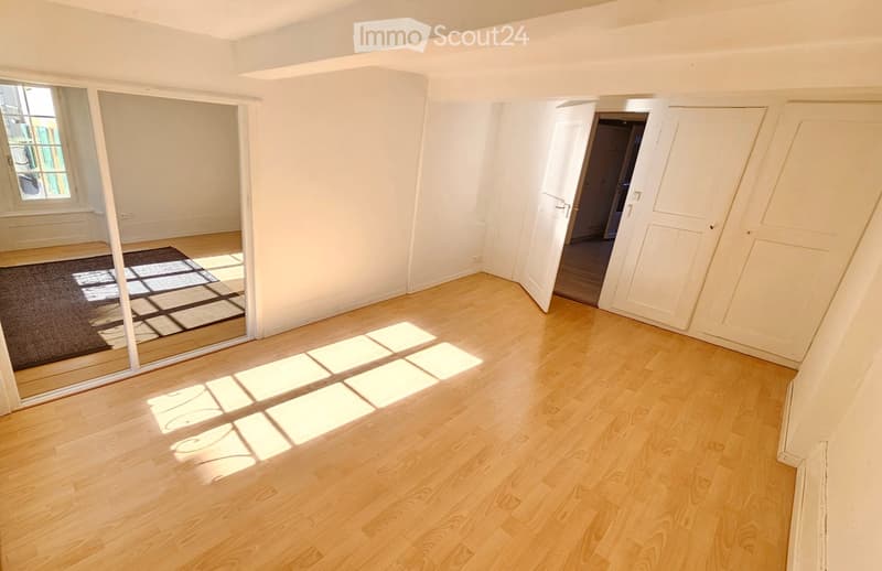 Empty room with wooden flooring, white walls, large window, and open doorway leading to another space.