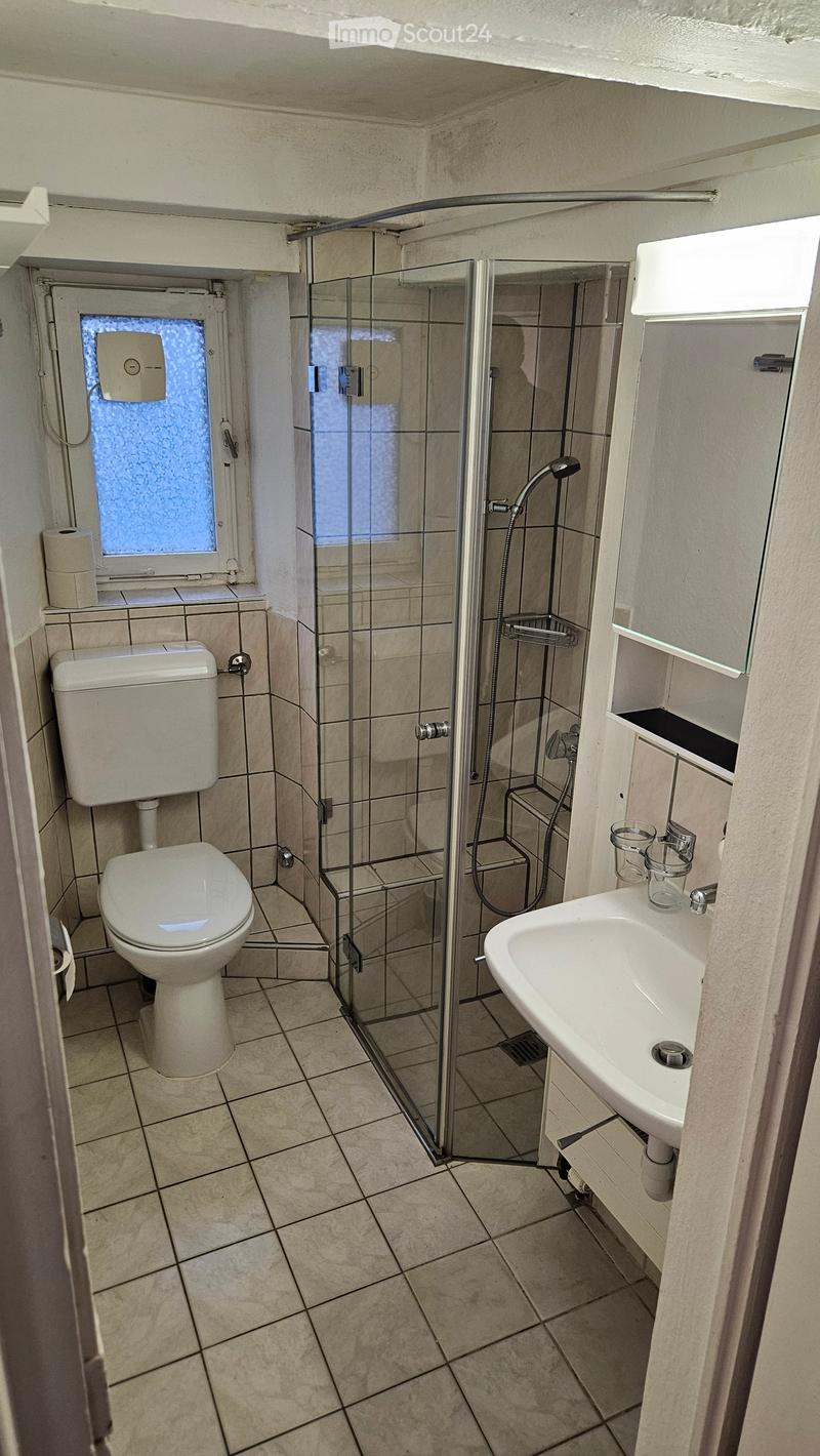 Modern bathroom with tiled walls and floor, featuring a toilet, sink, and glass-enclosed shower. Includes a window with frosted glass and a small shelf with cups.