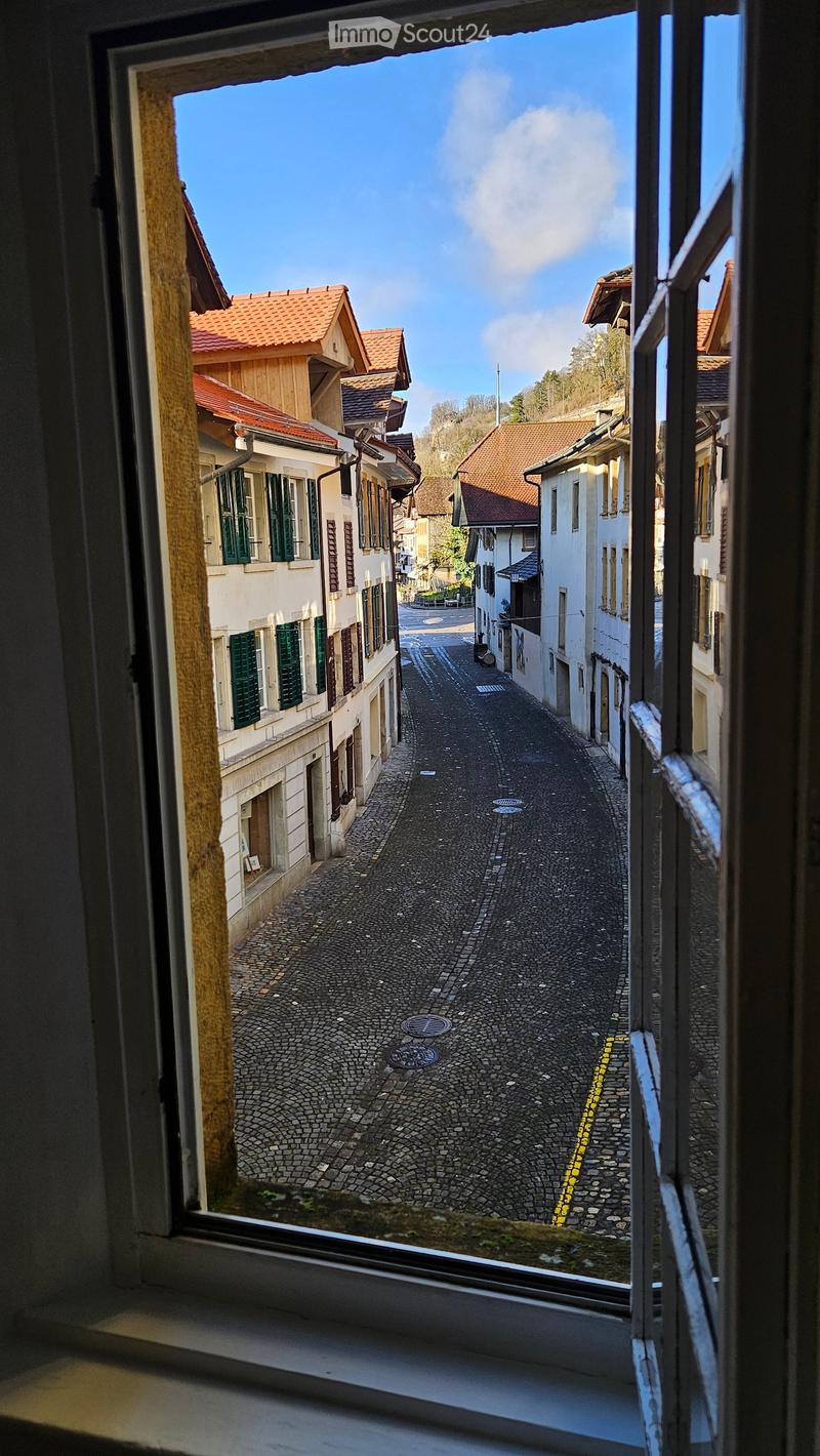 View from a window showing a narrow cobblestone street lined with traditional European houses featuring red-tiled roofs and green shutters.