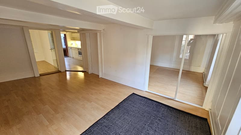 Empty room with wooden flooring, white walls, and large windows. A small black rug is placed on the floor. The room is connected to a kitchen area visible through an open doorway.