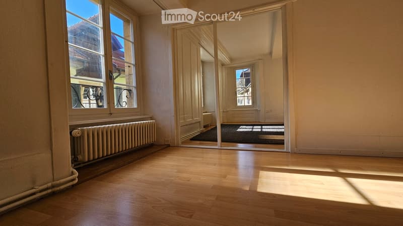 Empty room with wooden flooring, large windows, radiator, and a mirror reflecting another room.
