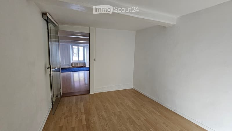 Empty room with wooden flooring and white walls, featuring a glass door leading to another room with a window and radiator.