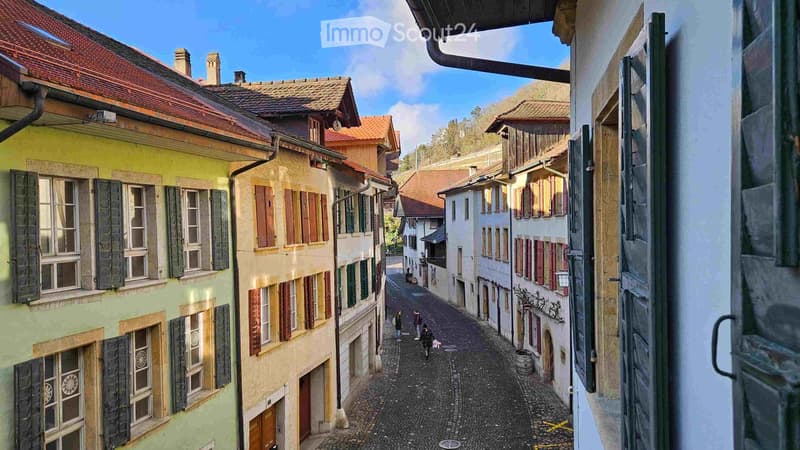 A narrow cobblestone street lined with colorful, multi-story buildings with traditional European architecture, featuring wooden shutters and tiled roofs.
