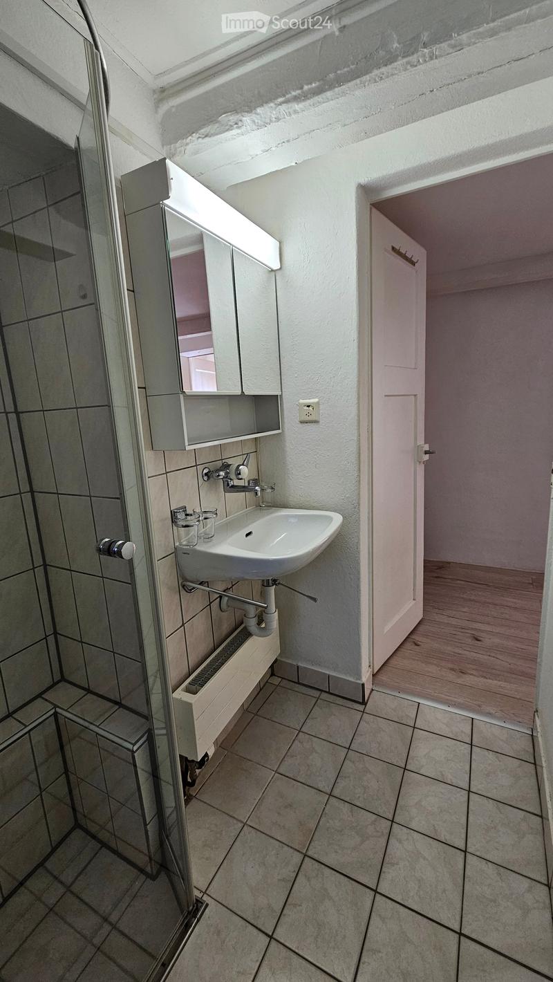 Bathroom with a white sink, mirror, and a glass shower enclosure. The floor is tiled, and there is a wooden door leading to another room.