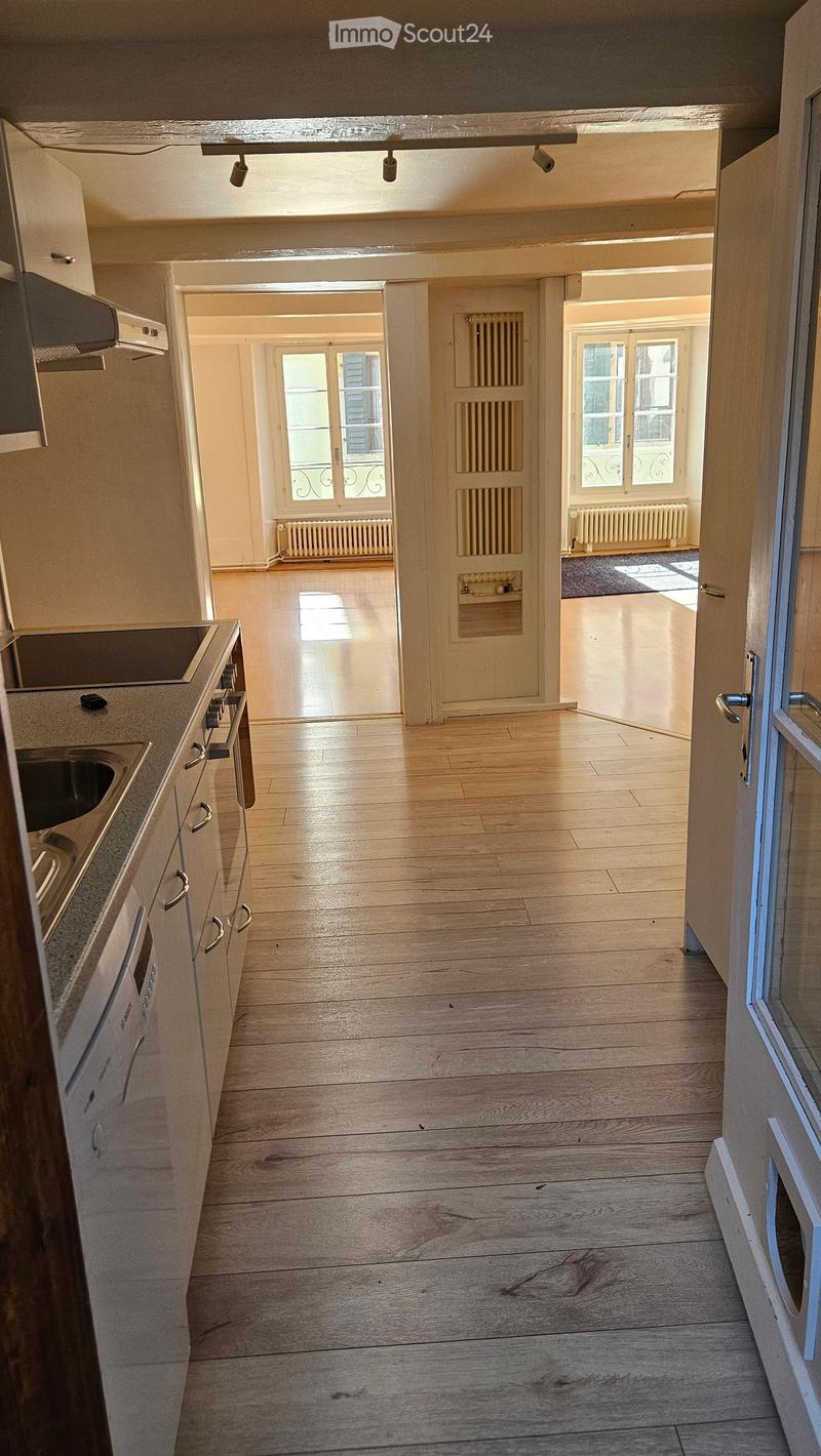 Bright hallway with wooden flooring, leading to a kitchen area on the left and an open living space on the right. The kitchen features white cabinets, a countertop, and built-in appliances.