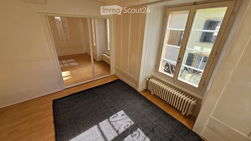 Empty room with wooden flooring, large window with view, radiator, and a small rug near the window.