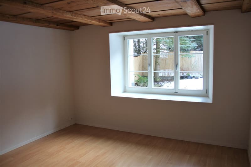 Empty room with wooden flooring, exposed wooden ceiling beams, and a large white-framed window showing a view of a snowy outdoor area with a fence and some plants.