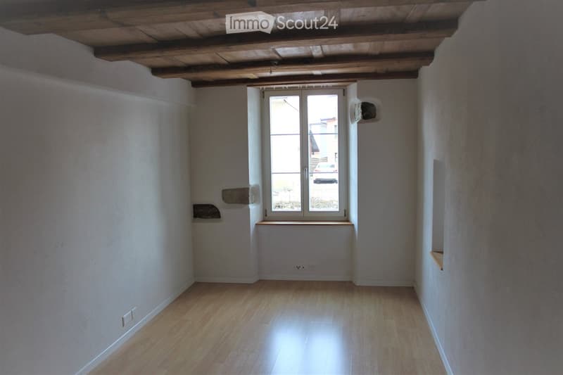 Empty room with wooden flooring, white walls, and a large window with a view of the outside. The ceiling has exposed wooden beams.