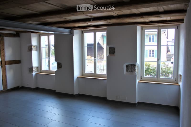 Empty room with exposed wooden ceiling beams, tiled floor, and large windows with wooden sills. The walls are white with some exposed stone patches.