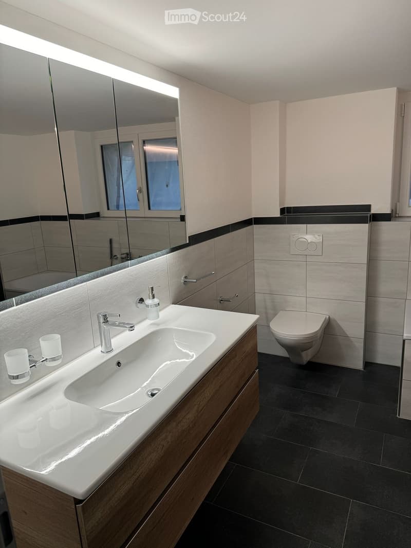 Modern bathroom with a white sink, chrome faucet, wooden vanity, toilet, large mirror with built-in lighting, and two decorative cups on the sink.