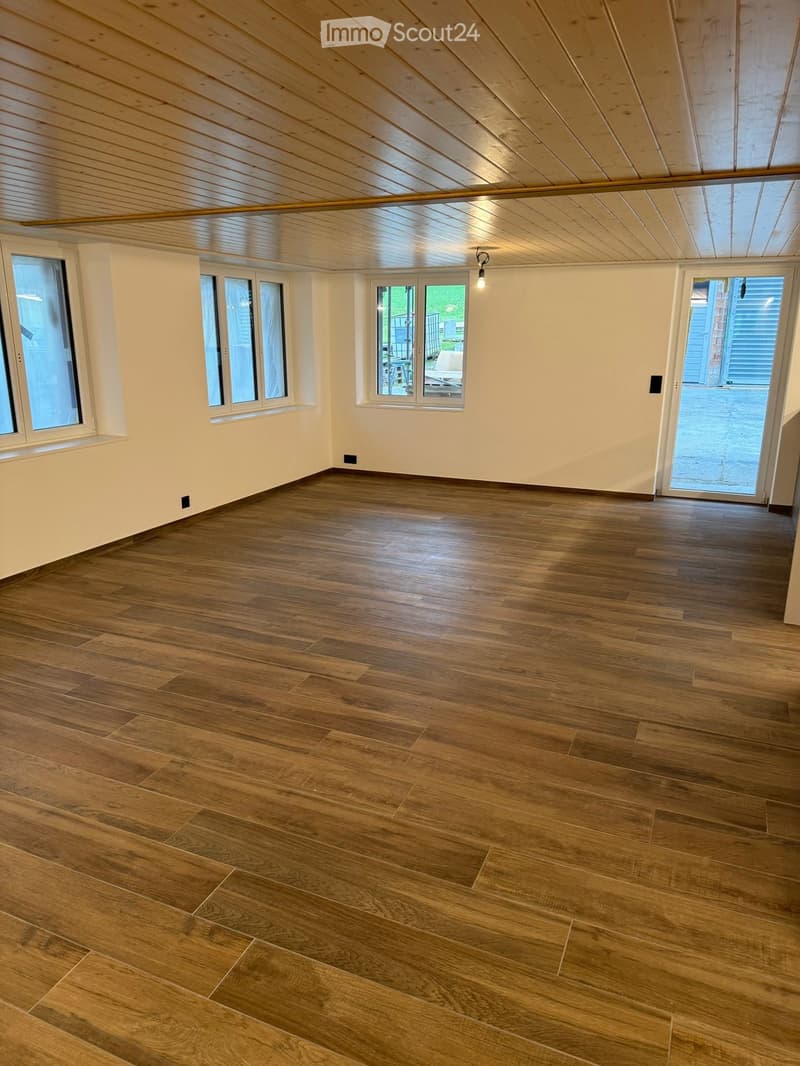 Empty room with wooden flooring, white walls, large windows, and a sliding glass door leading outside.
