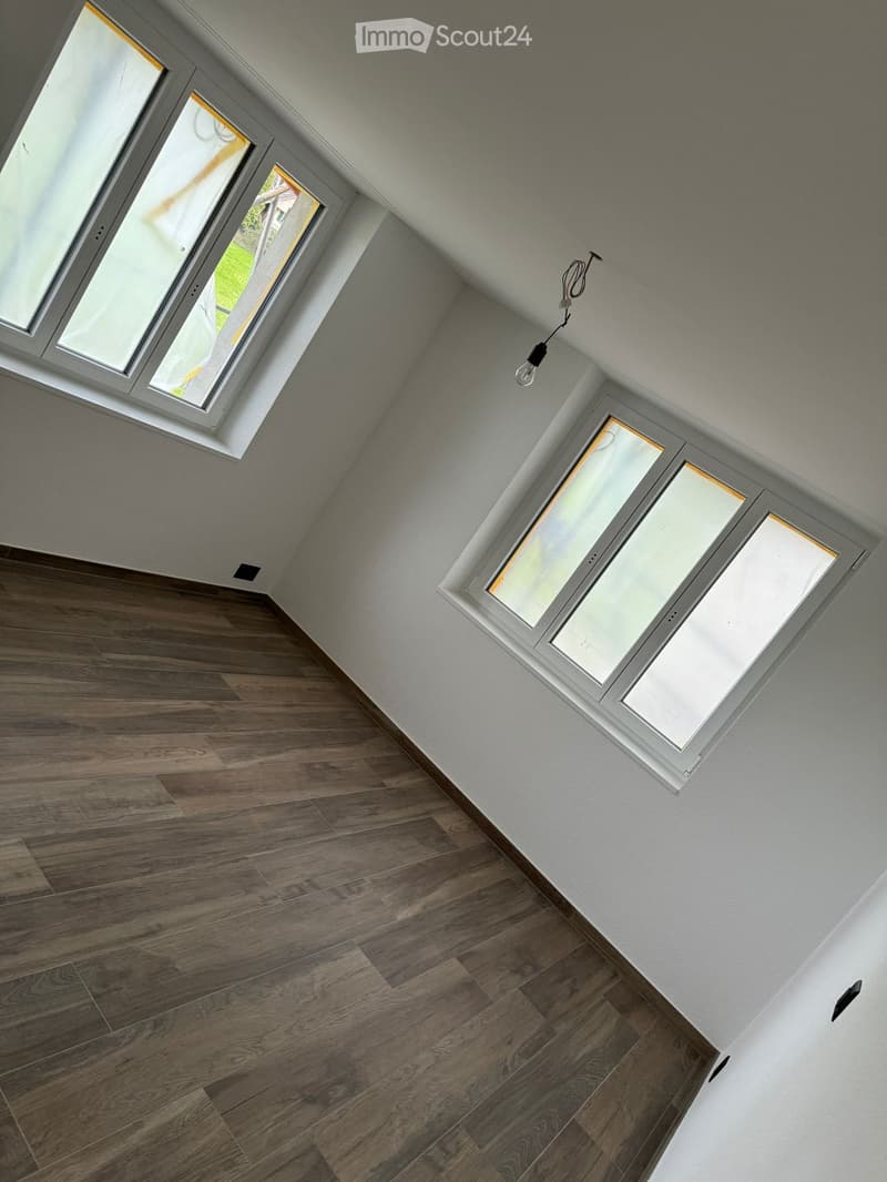 Empty room with wooden flooring and white walls, featuring multiple windows with white frames and a single light fixture hanging from the ceiling.