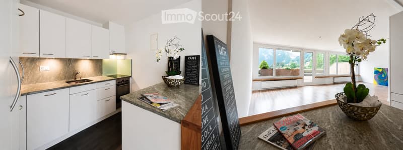 Modern kitchen with white cabinets, granite countertop, and a large open living area with a view of the mountains. The living area features a large window, a vase with white flowers, and a table with magazines.