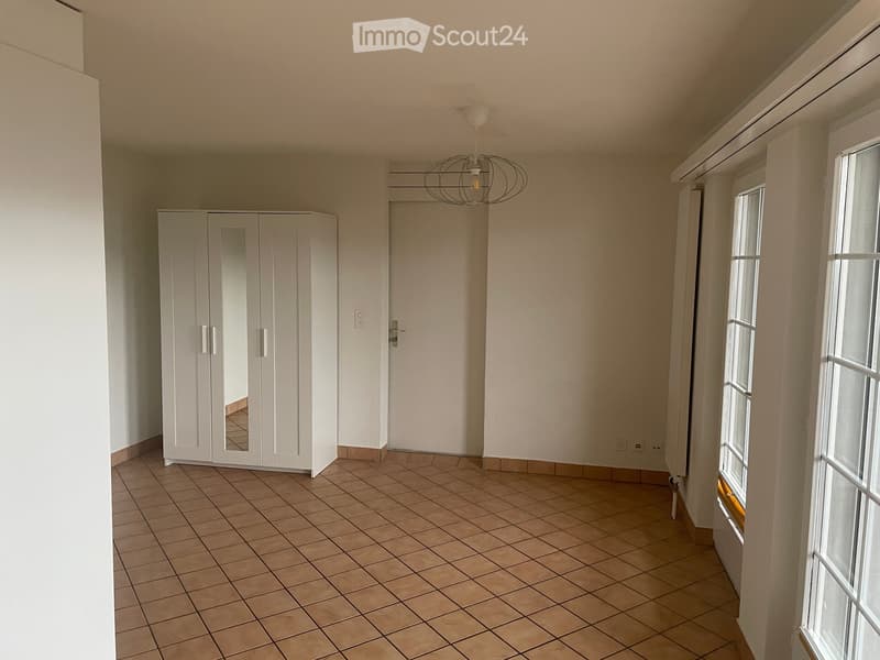 Empty room with tiled floor, white walls, large windows, and a white wardrobe with mirrored doors.