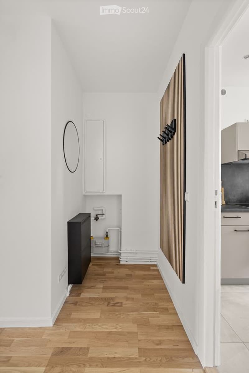 A narrow hallway with light wooden flooring, white walls, and a small built-in cabinet. A round mirror is mounted on the left wall, and a coat rack is on the right. The hallway leads to a kitchen area visible at the far end.
