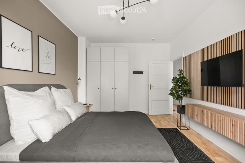 Modern bedroom with a grey bed, white pillows, a nightstand, framed artwork, a potted plant, a cabinet, and a TV mounted on a wooden wall.