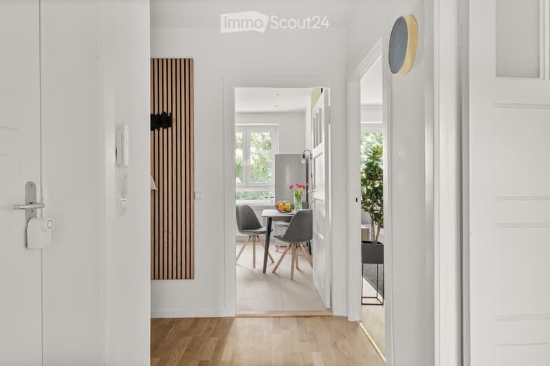 A bright, modern hallway with white walls and wooden flooring, leading to a small dining area with a table, chairs, and a vase of flowers. A potted plant is visible in the adjacent room.
