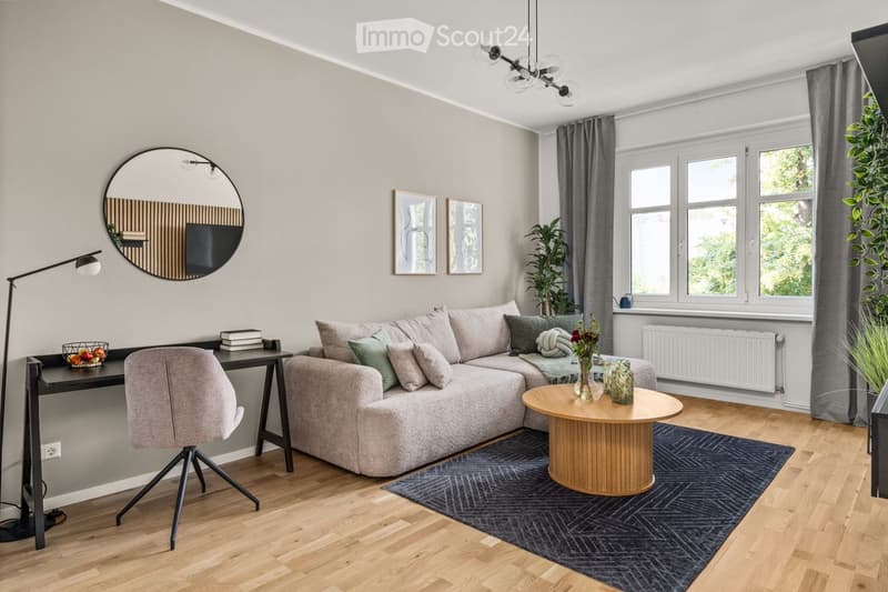 Modern living room with a grey sectional sofa, wooden coffee table, grey armchair, round mirror, floor lamp, and decorative items on a side table.