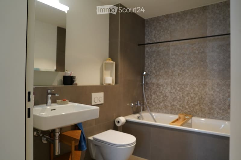 Modern bathroom with a white sink, chrome faucet, toilet, bathtub, and a small wooden stool holding a towel. The walls are tiled in a neutral color scheme, and there is a mirror above the sink.
