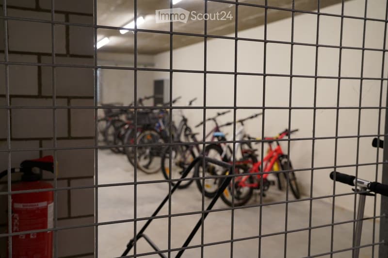 A view through a metal grid shows a parking area with several bicycles lined up, a red fire extinguisher on the left, and a concrete floor.