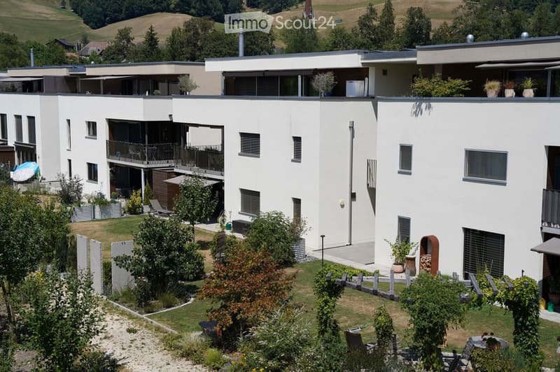 Modern two-story residential building with white exterior, multiple balconies, and surrounding garden area with patio furniture and greenery.