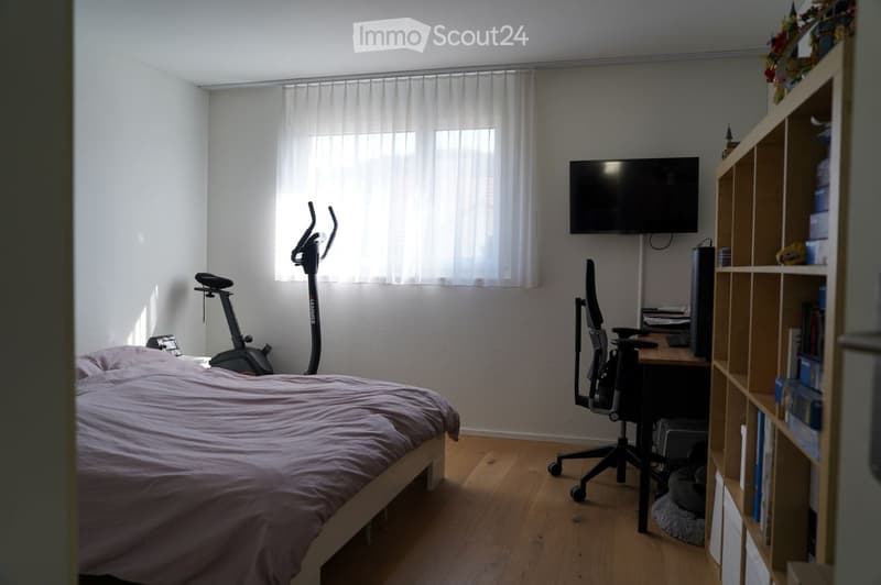 A small, well-lit bedroom with a bed, a desk with a chair, a mounted screen, an exercise bike, and a wooden shelf with books and decorative items.