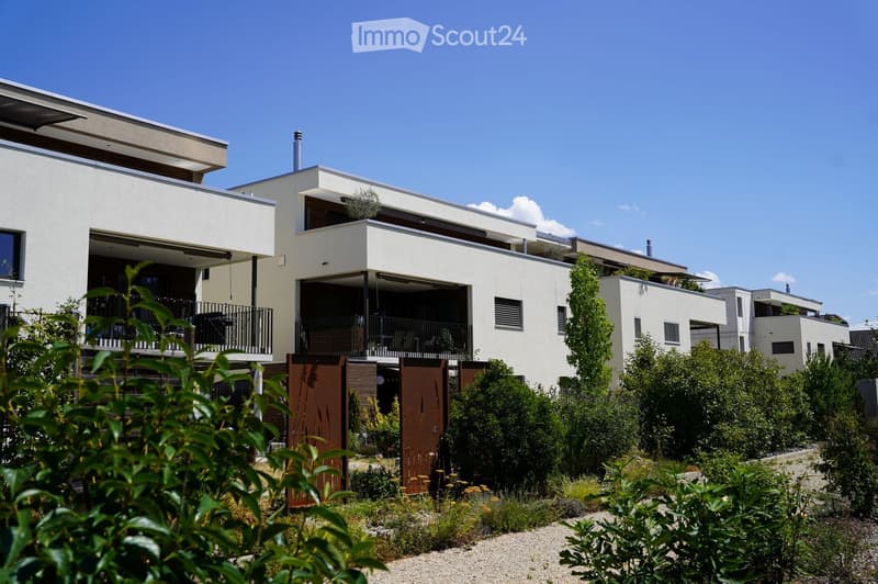 Modern two-story house with white exterior, large windows, and a balcony on the upper level. The house features a flat roof and contemporary design, surrounded by greenery and a small garden area.