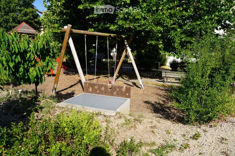 A playground area with a wooden swing set, surrounded by trees and greenery, with a small wooden playhouse and a bench nearby.