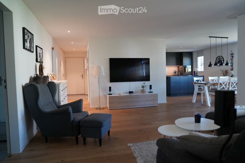 Modern living and dining area with wooden flooring, grey sofa, armchair, ottoman, coffee table, TV, dining table with chairs, and kitchen area with cabinets and pendant lights.