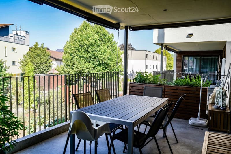 Balcony with a wooden table and several chairs, surrounded by greenery and a metal railing, overlooking a cityscape with buildings and trees.
