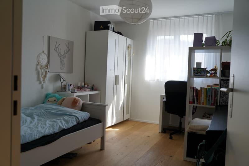 Bright bedroom with wooden floor, bed with blue blanket, desk with chair, bookshelf, wardrobe, and decorative items including a dreamcatcher and picture frame.