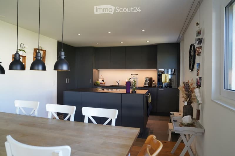 Modern kitchen with dark grey cabinets, a central island, and a dining area with a wooden table and white chairs. The space features pendant lighting, a stainless steel sink, and various appliances.