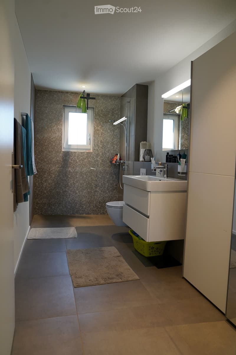 Modern bathroom with a white sink, toilet, large mirror, and tiled walls. Includes towels, a small rug, and a shower area with a glass enclosure.