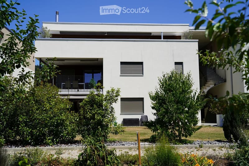 Modern two-story house with white exterior, large windows, and a balcony on the upper level. The house is surrounded by a well-maintained garden with various plants and shrubs.