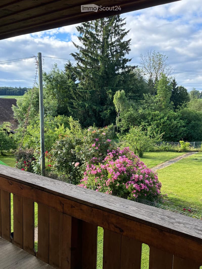 Wooden balcony overlooking a lush garden with pink flowers, green grass, and tall trees under a partly cloudy sky.