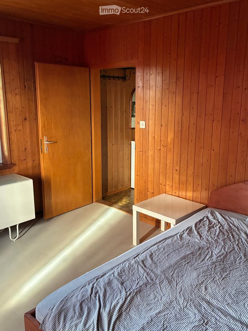 Cozy bedroom with wooden walls and flooring, featuring a bed with striped bedding, a small nightstand, a table, and a cabinet.