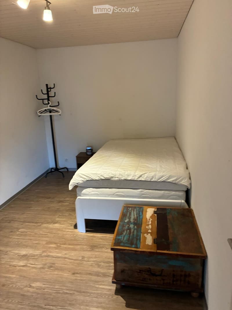 Small bedroom with a single bed, nightstand, and coat rack. Light wooden flooring and white walls.