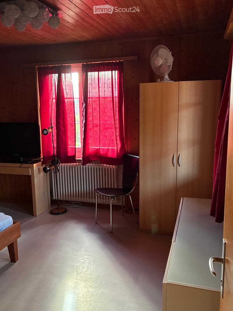 Cozy bedroom with wooden walls, a bed, a chair, a wardrobe, a fan on top, a table with a lamp, and red curtains on the window.