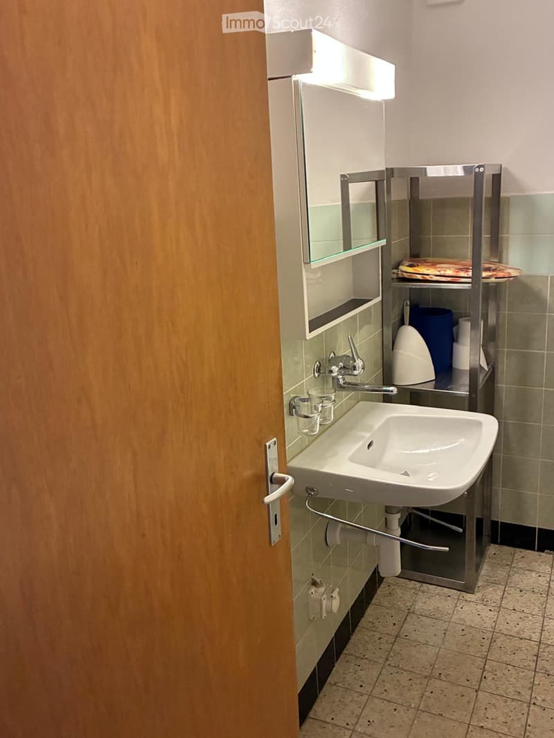Modern bathroom with a white sink, chrome faucet, mirror cabinet, and a toilet in the corner. The walls are tiled in a light green color, and the floor has beige tiles.