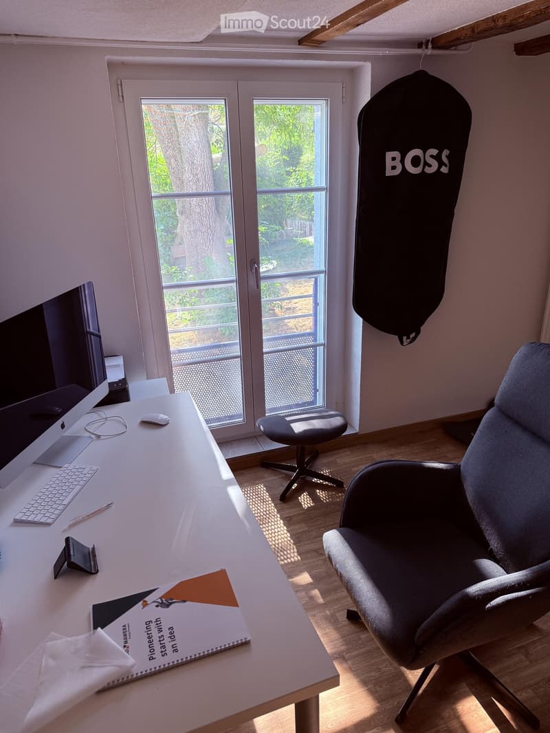 A modern office space with a large desk, ergonomic chair, laptop, keyboard, mouse, and a large window with a view of greenery.