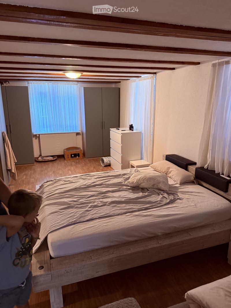 Cozy bedroom with wooden bed, nightstand, drawer unit, cat bed, and cat tree. Light-colored bedding and curtains, wooden floor, and exposed wooden beams on the ceiling.