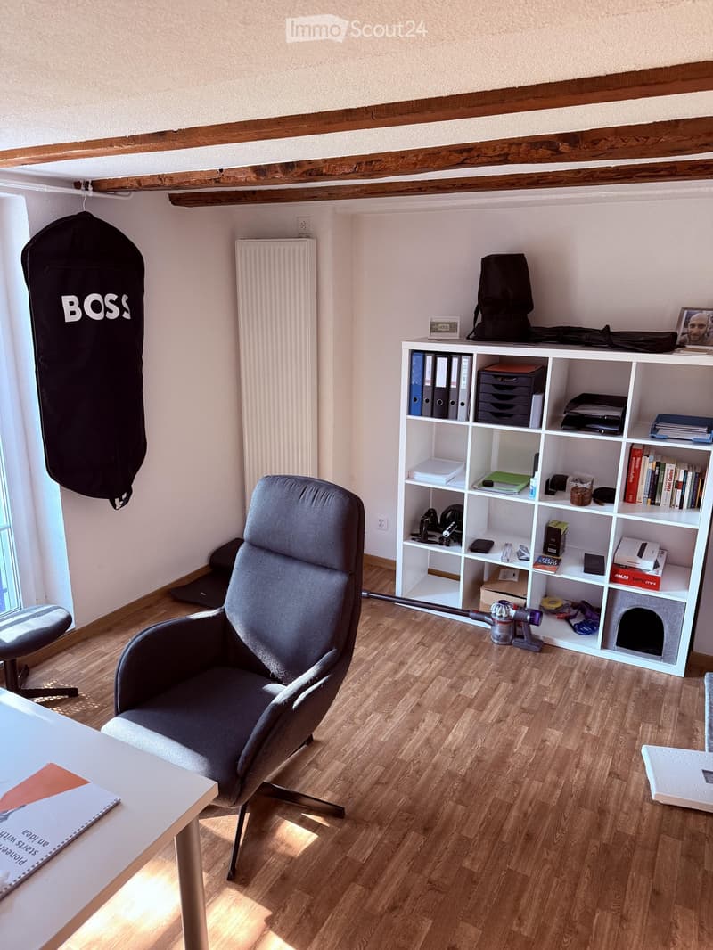 A well-lit room with wooden flooring, a large grey armchair, a white table, a white shelving unit filled with books and office supplies, a radiator, a backpack, a laptop, a cat house, and a vacuum cleaner.