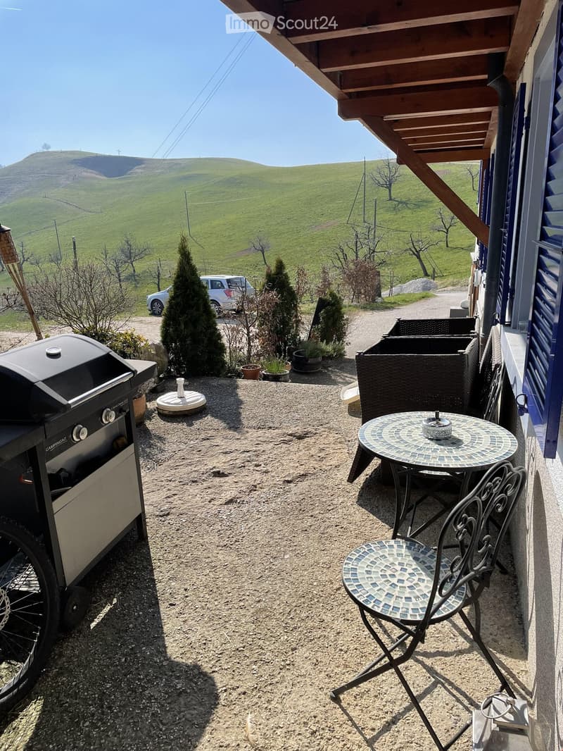 Outdoor patio area with a round mosaic table, matching chair, grill, and wicker basket, overlooking a grassy hillside.