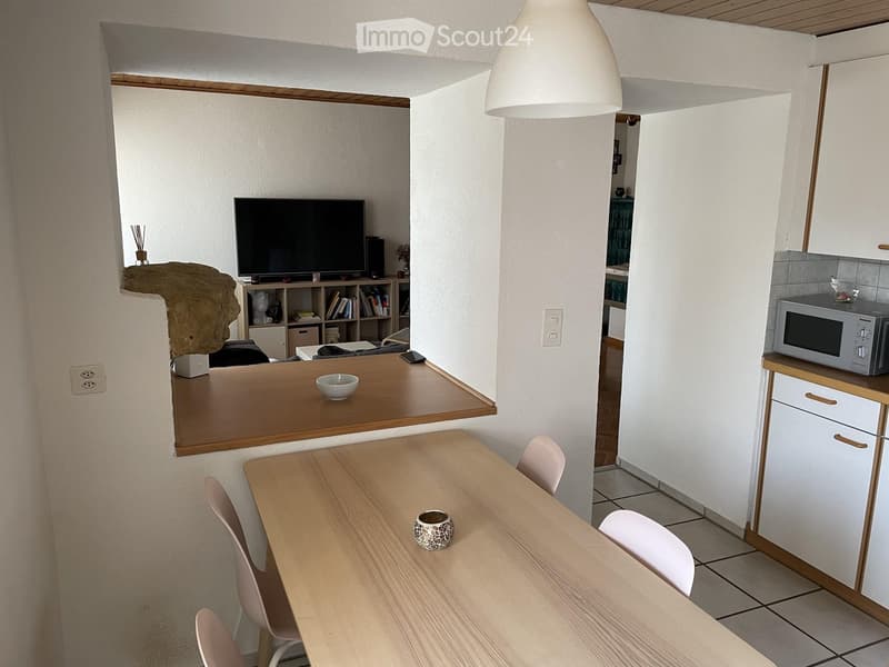 A modern, open-plan living and dining area with a wooden dining table, chairs, a microwave, a television, and a shelf with books and decorative items.