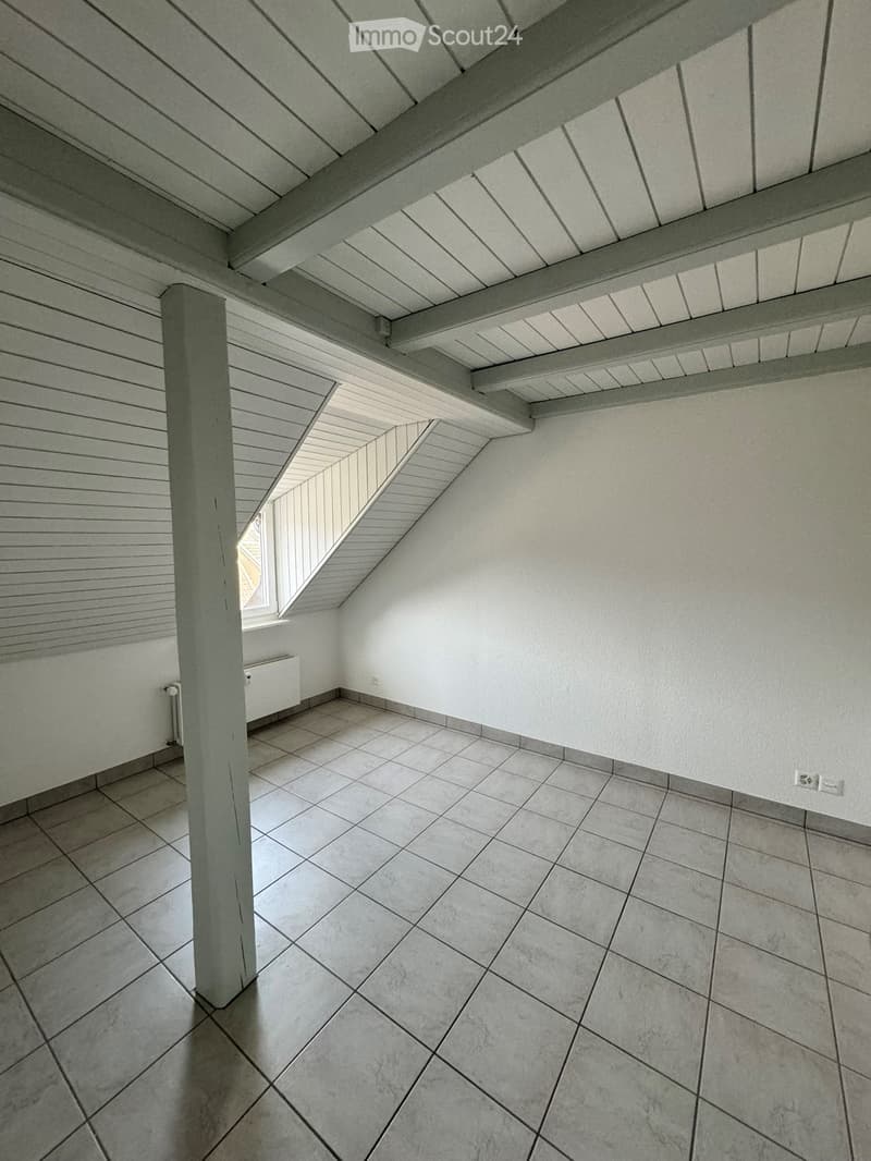 Empty attic space with slanted wooden ceiling, tiled floor, and a small window letting in natural light.