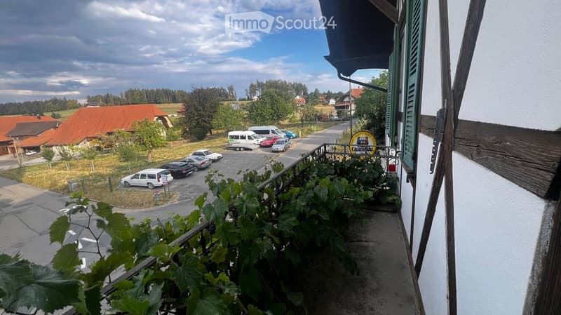 Balcony overlooking a rural road with parked cars, houses, and greenery in the background.