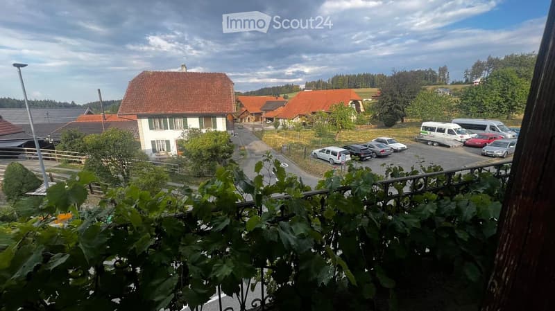 Balcony view overlooking a rural village with traditional houses, red-tiled roofs, parked cars, and greenery.