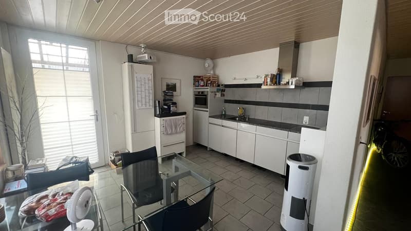 Modern kitchen and dining area with white cabinets, stainless steel appliances, a glass dining table with chairs, and a fan. The space features tiled flooring and large windows with blinds.