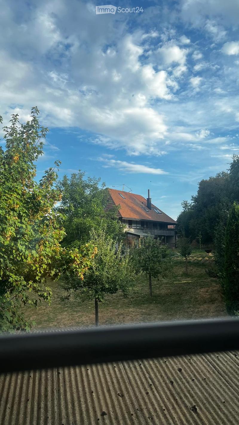 A two-story house with a red-tiled roof and a chimney, surrounded by trees and greenery under a partly cloudy sky.